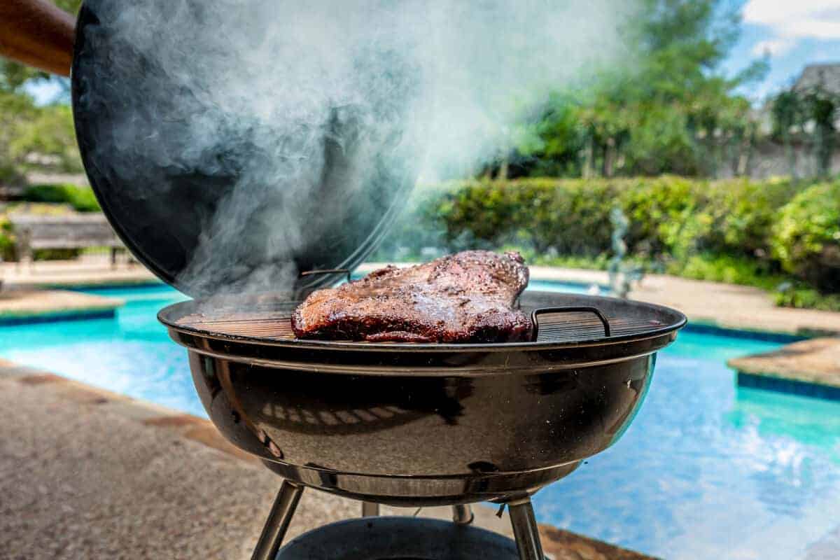 A large brisket on a kettle grill in front of a swimming pool