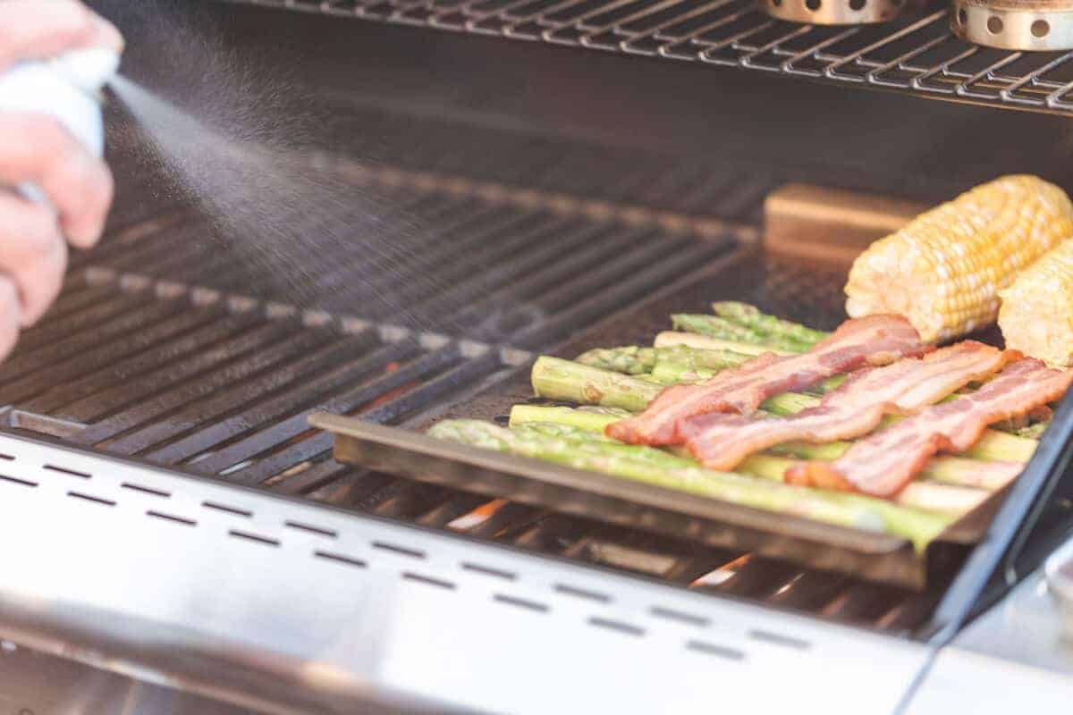 Cooking spray being sprayed onto bacon and asparagus on a gr.