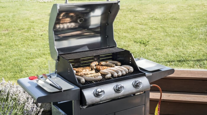 A 3 burner gas grill full of sausages and meats against a grass bg.