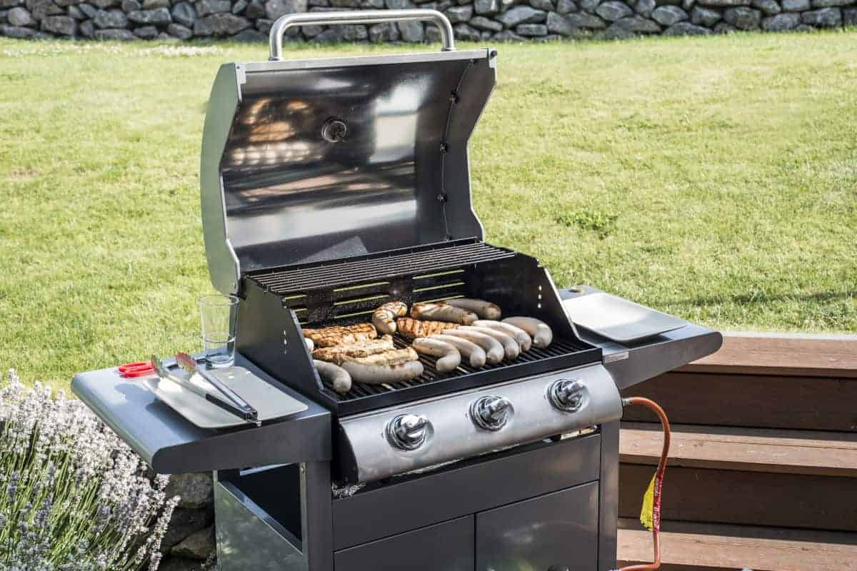 A 3 burner gas grill full of sausages and meats against a grass bg