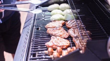 Gas grill with onions, teak and other bits, and a man holding tongs to flip them.