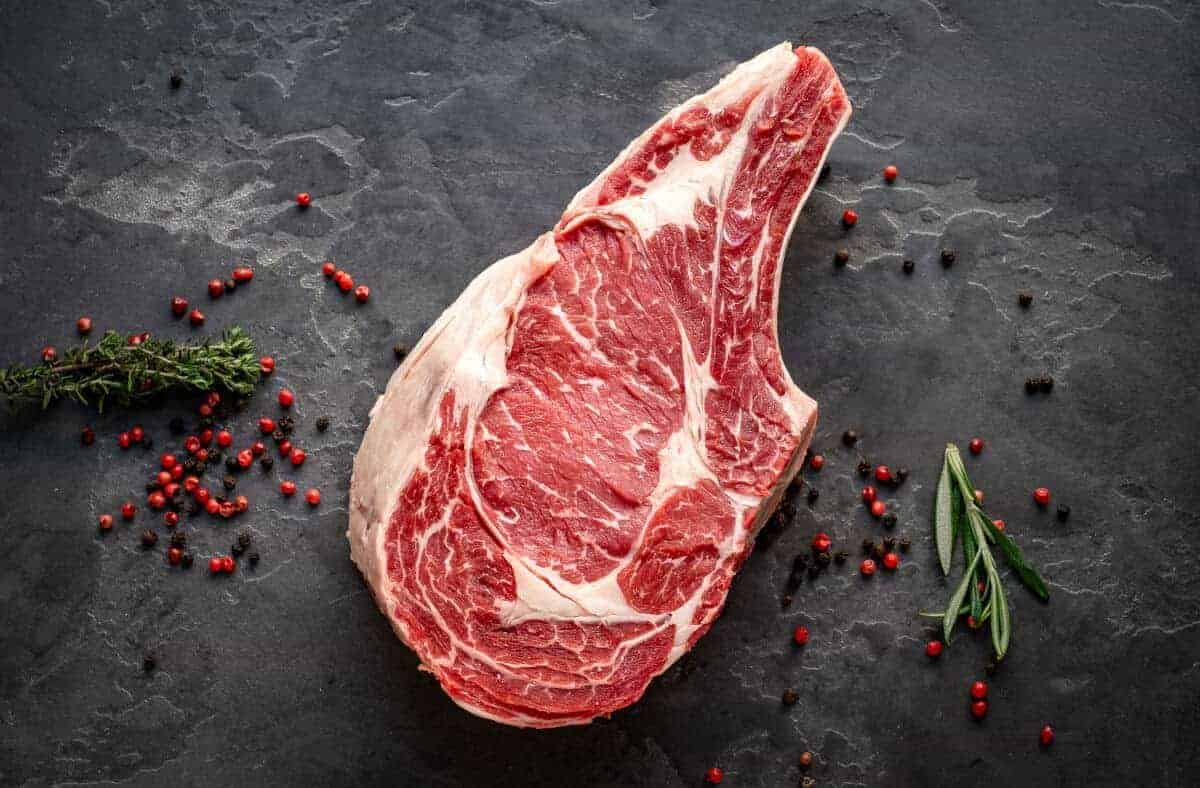 A bone-in ribeye cowboy steak on a dark surface with red peppercorns