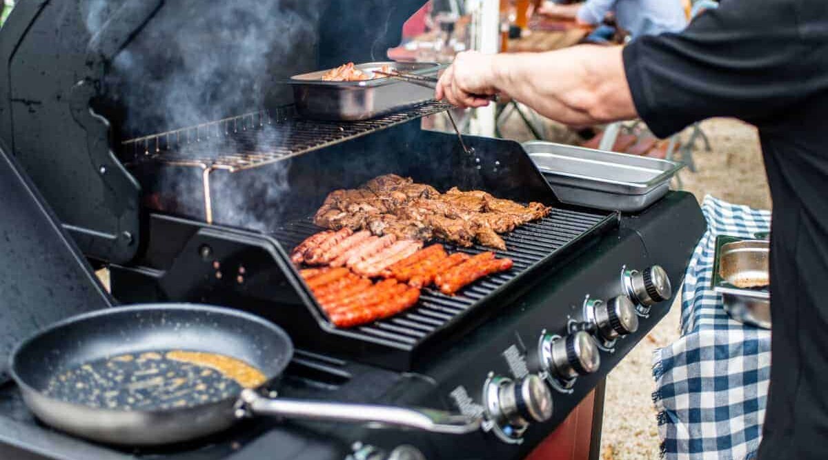 A gas grill with frying pan, bratwurst and steaks cooking.