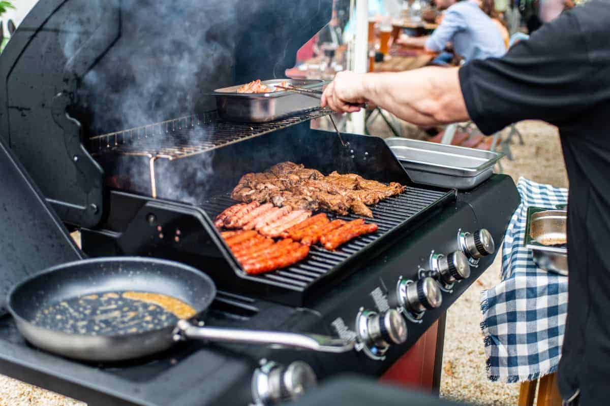 A gas grill with frying pan, bratwurst and steaks cook.