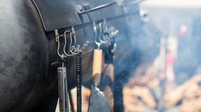 Close up of a barrel shaped smoker with utensils hanging off the front.
