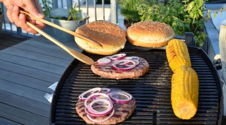 A couple of burgers and corn being grilled on a small grill on a balcony.