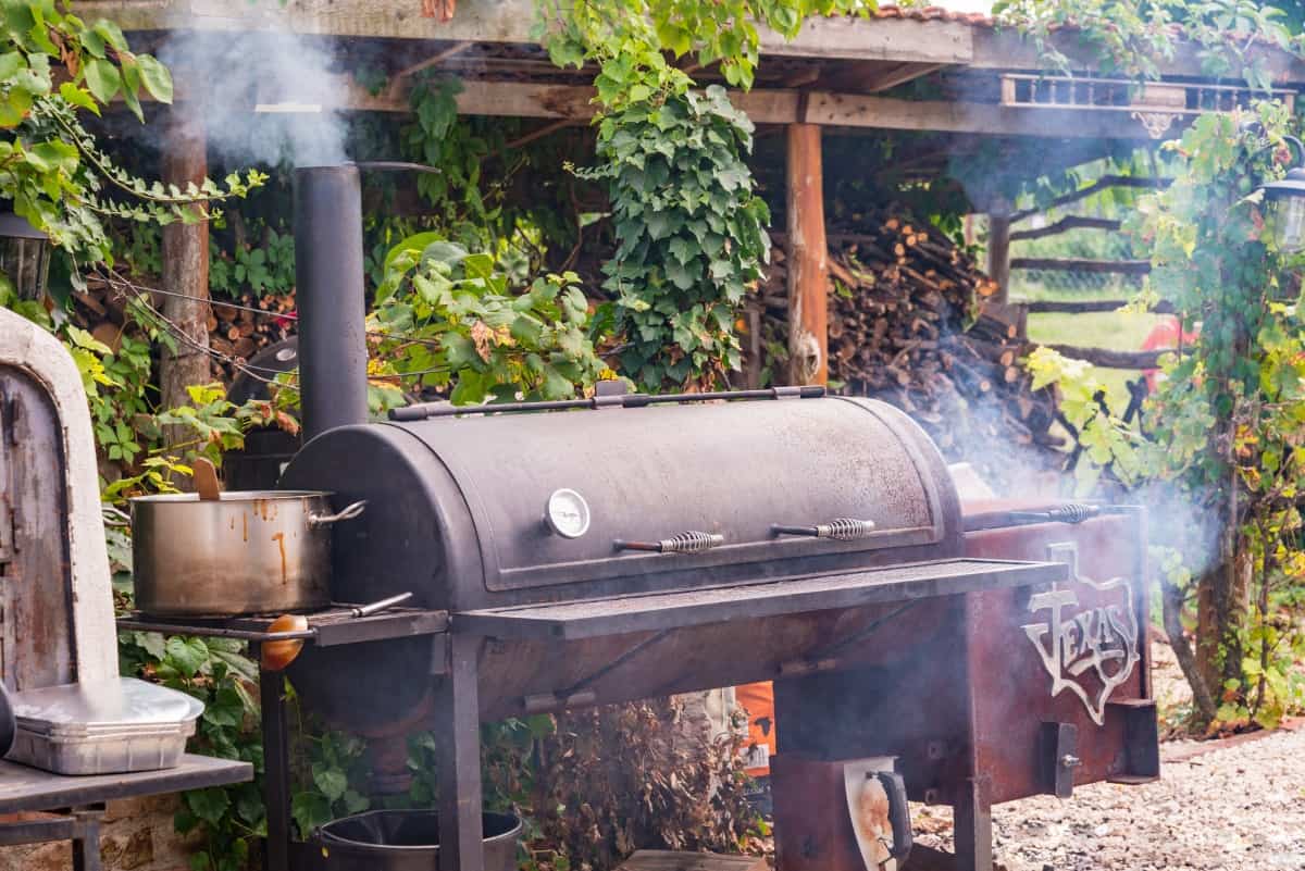 A well used offset smoker in a backyard in front of a log store.