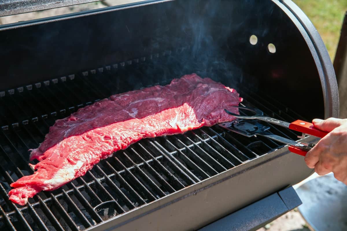 A large bavette steak being placed into a drum style grill with tongs