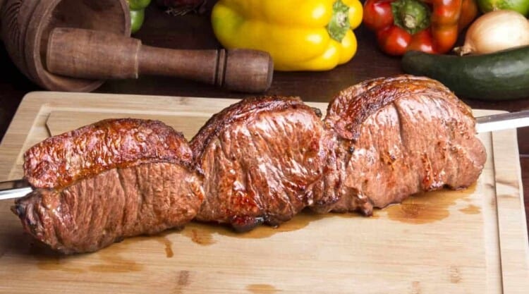 Skewered picanha steak on a chopping board with veg in background.