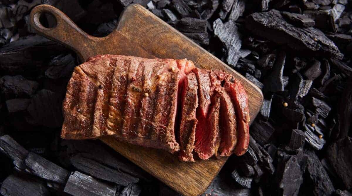 Cooked ranch steak, sliced on a chopping board sitting over charcoal.