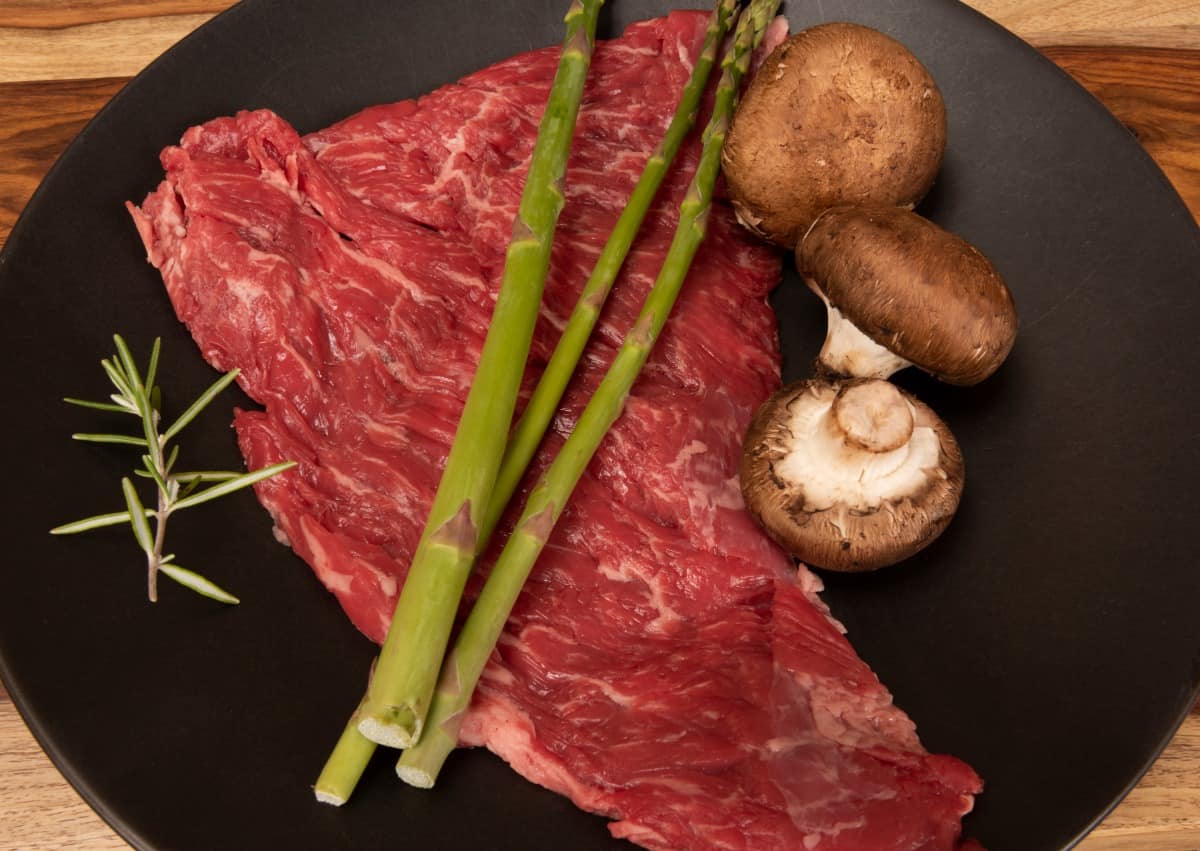 raw bavette steak on black plate with raw asparagus and mushro.