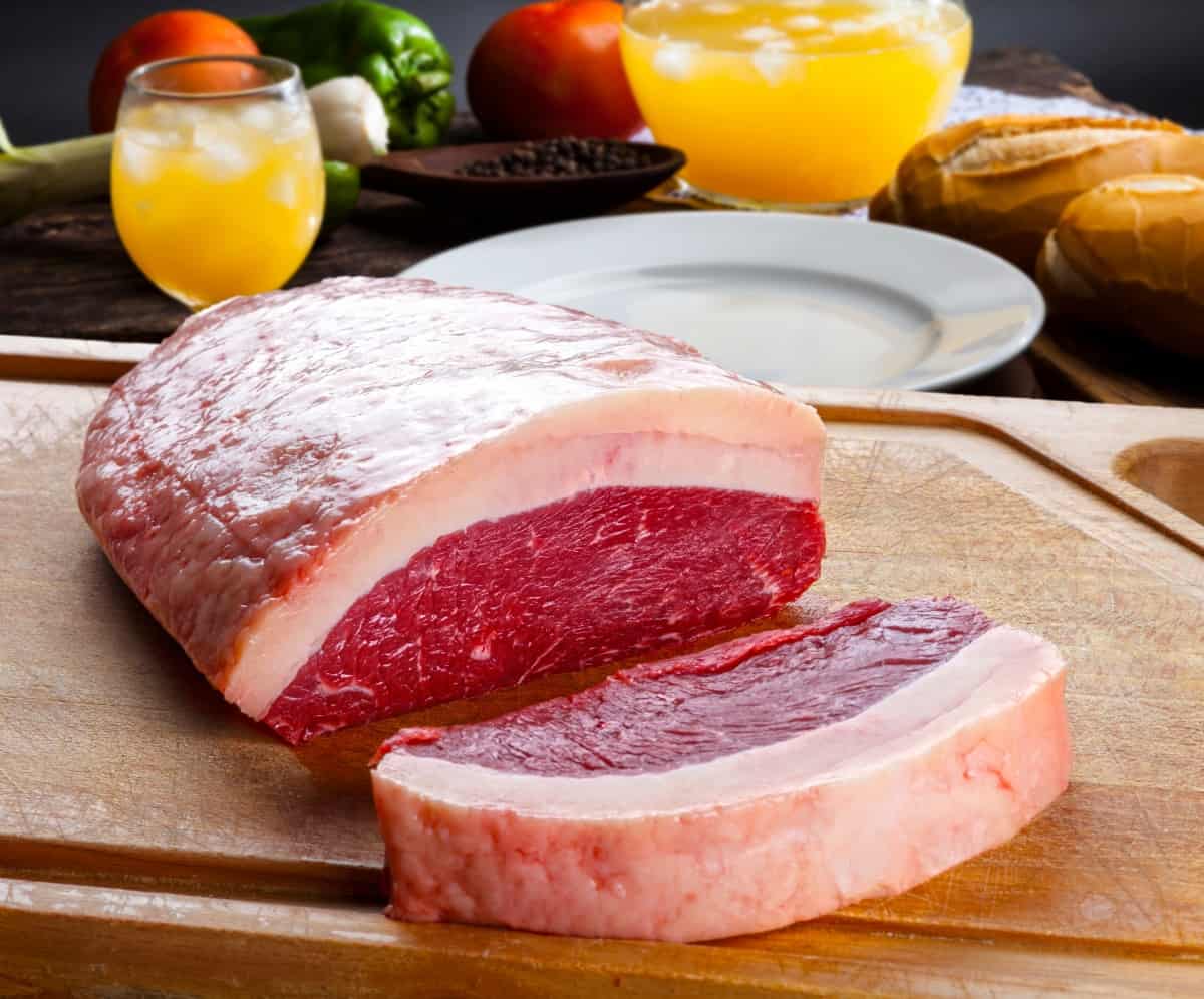 raw picanha joint with a steak sliced off, sitting on a cutting board in front of blurred vegetable backgro.