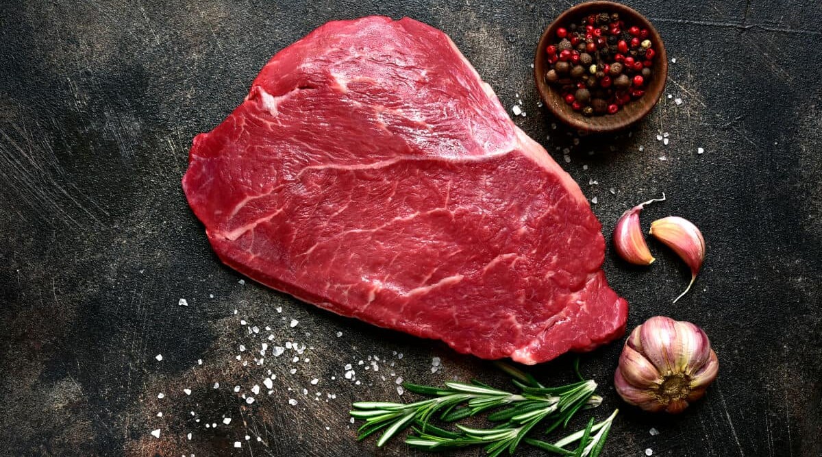 A raw shoulder steak on a dark surface with rosemary sprigs.