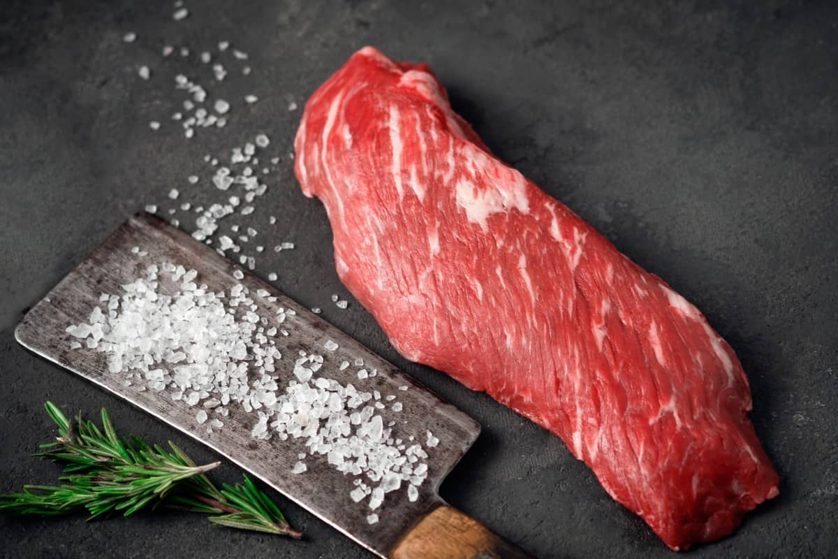 A raw skirt steak next to a meat cleaver covered with s.