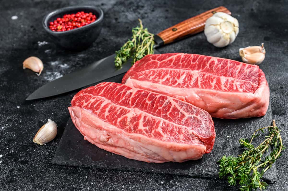 Two raw top blade steaks on a slate board, with garlic, pink peppercorns and th.