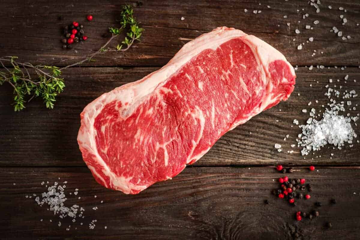 Raw sirloin steak on wood table, surrounded by salt pepper and he.