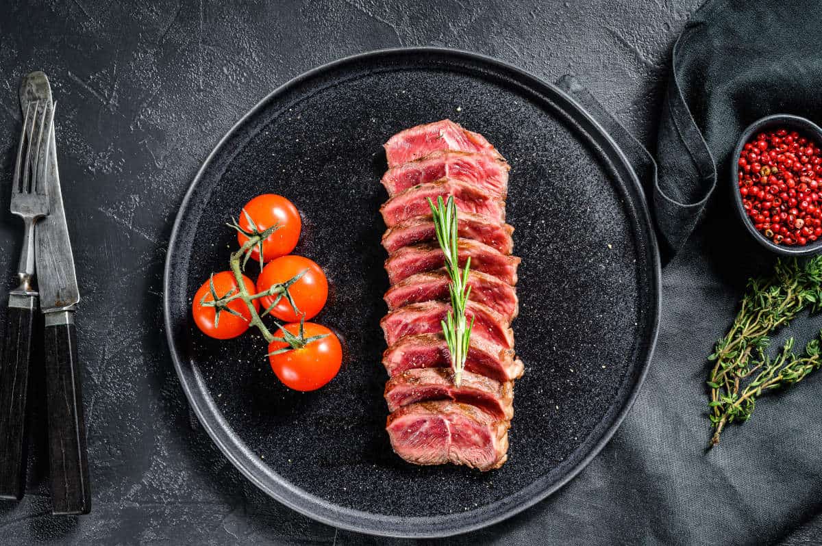 sliced grilled top blade steak on a black plate with some cherry tomat.