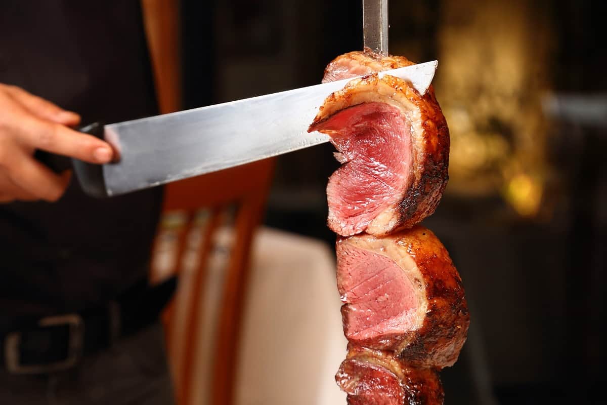 Skewered, grilled picanha being sliced with a large carving kn.