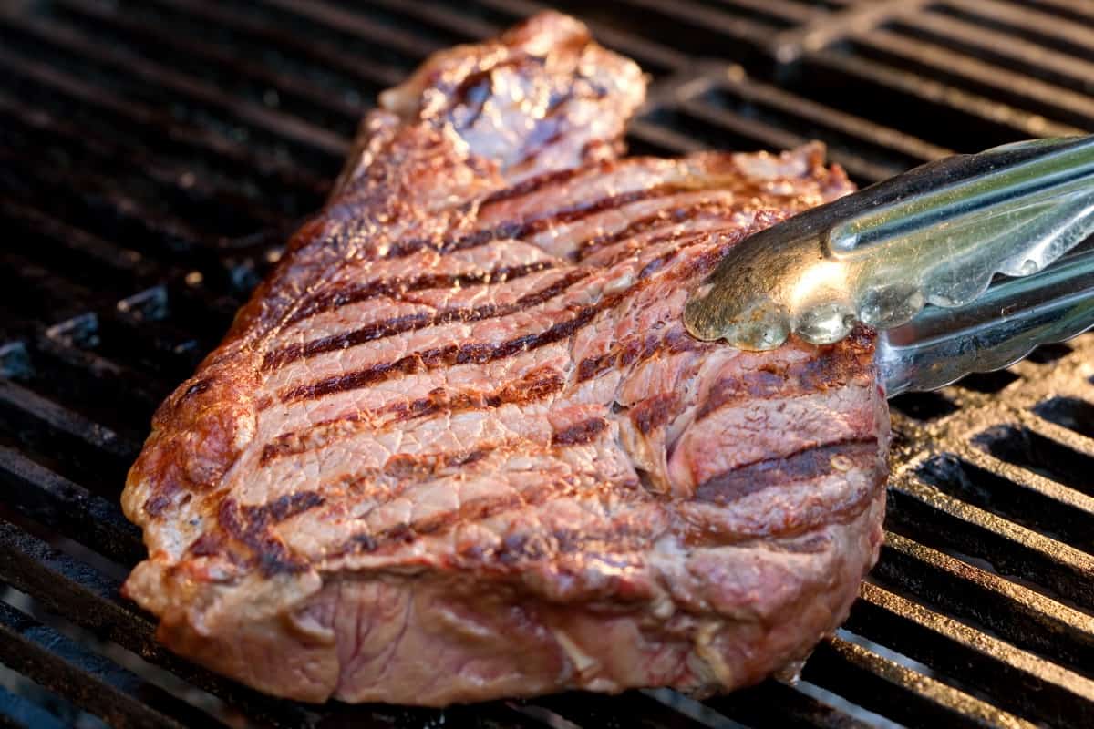 steak held in tongs over the grates of a pellet gr.