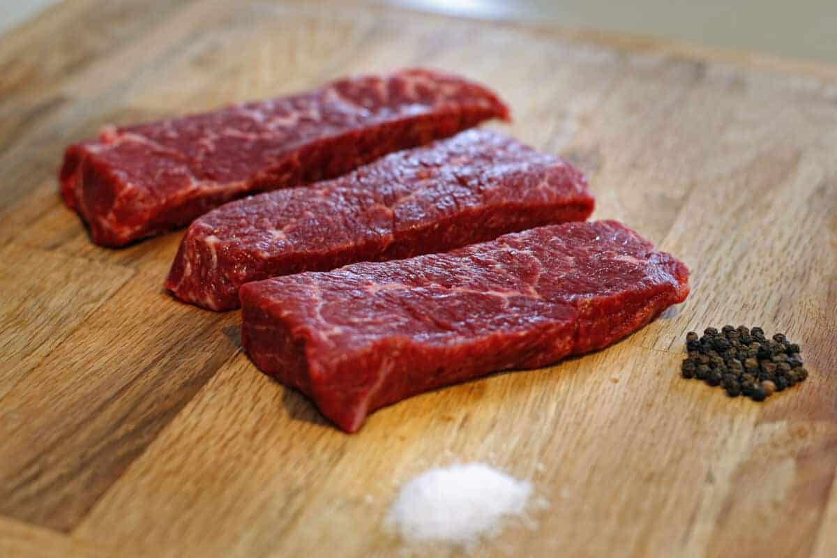 3 raw Denver steaks on a wooden surface with salt and pepper pi.