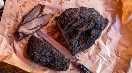A brisket slicing knife sat between a halved and sliced brisket on pink butcher paper.