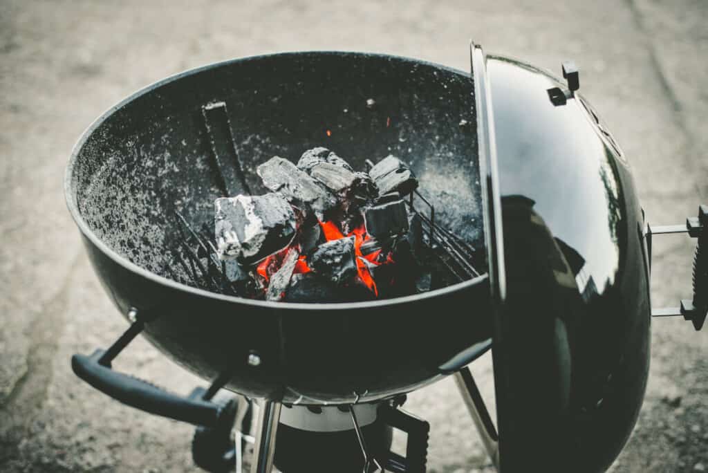 A lit, circle shaped charcoal grill with the lid hanging from the s.