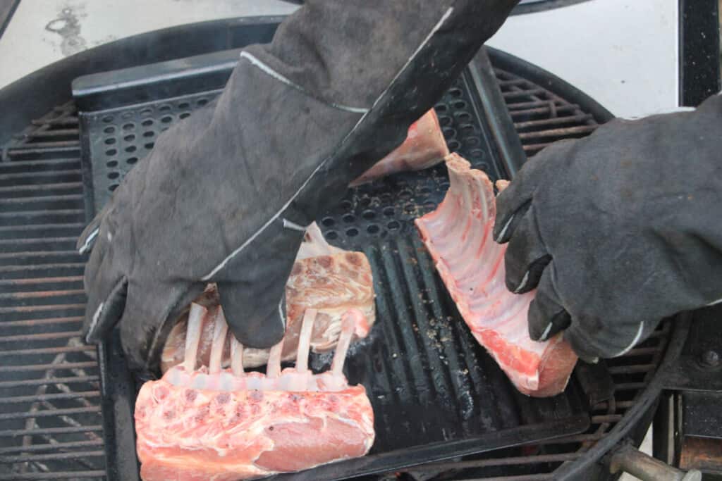Gloved hands moving lamb racks about a hot gr.