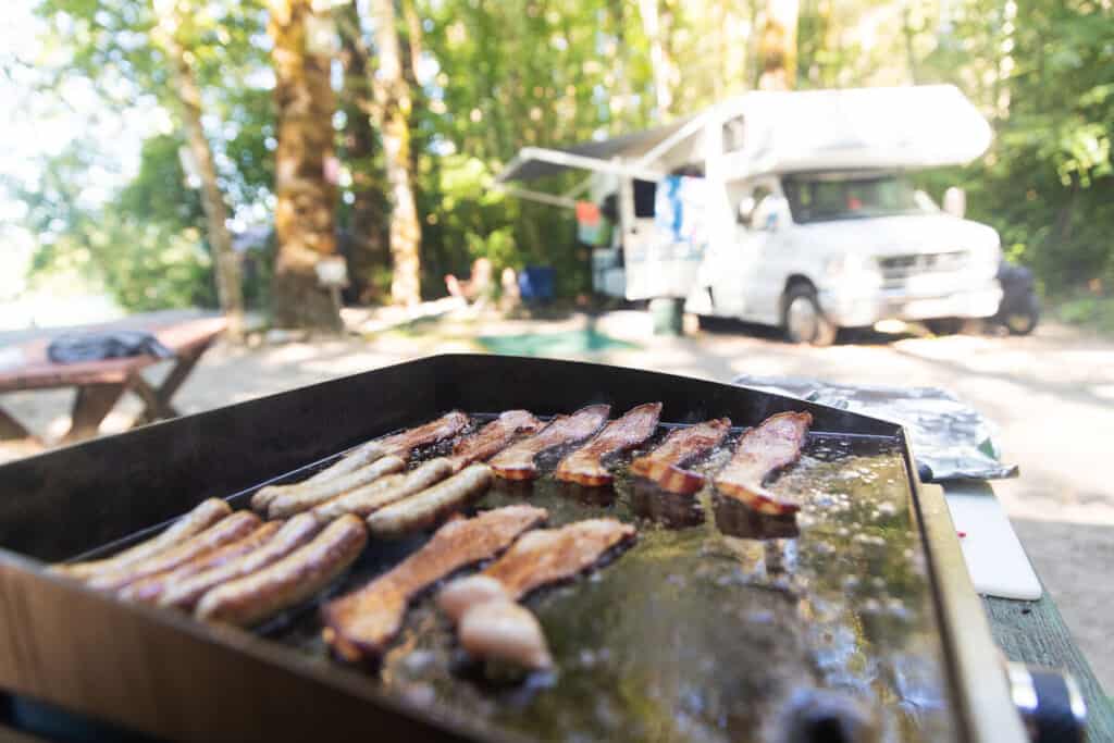 A portable flat top grill cooking bacon, with an RV in the backgro.