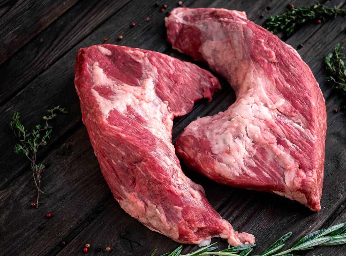 Two raw tri-tip on a wooden table surrounded by rosemary and th.