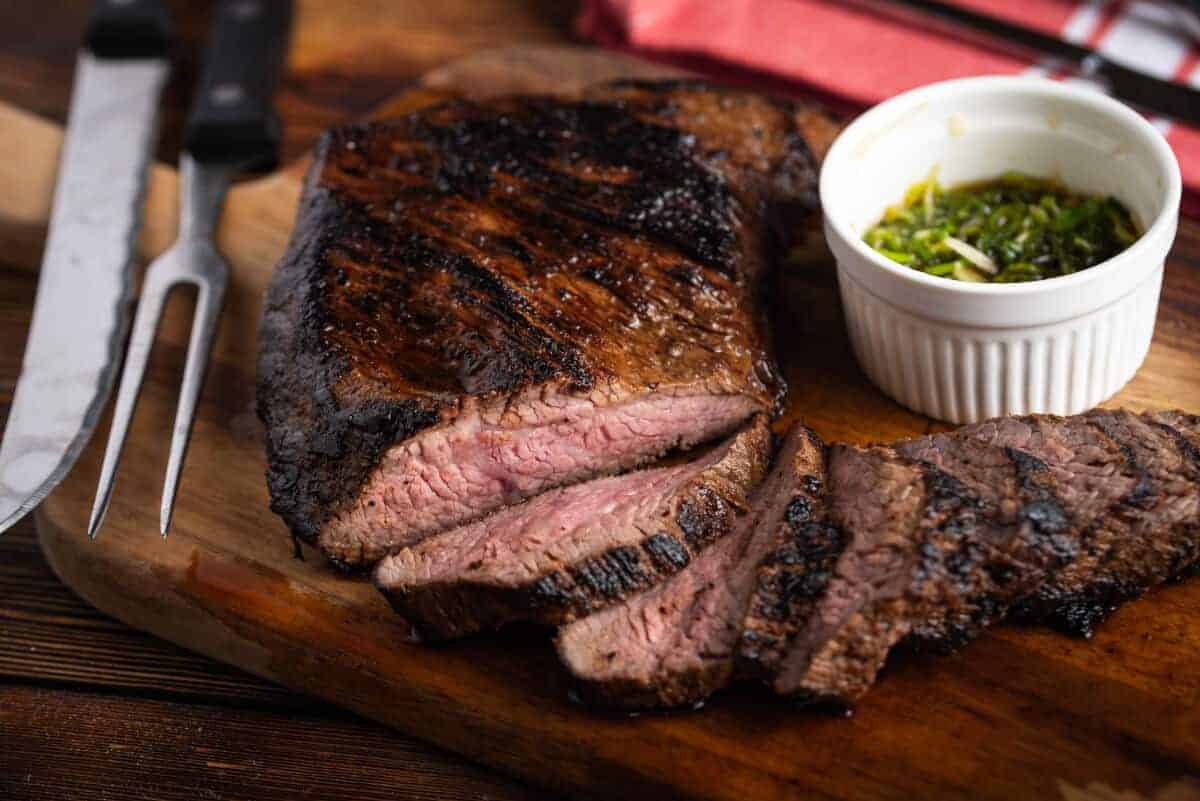 Sliced tri-tip, on a cutting board with a knife and ramekin of chimichu.