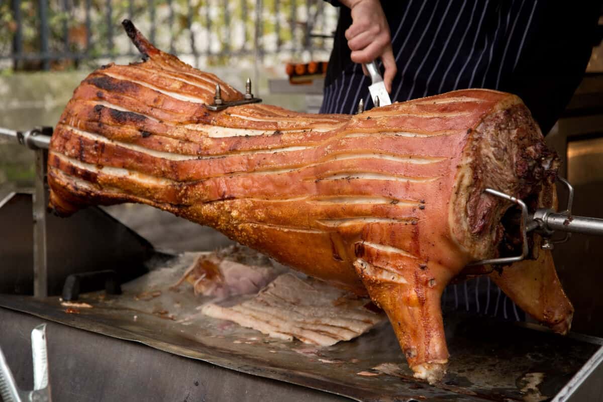 A whole hog on a rotisserie, with scored skin and a man about to slice.