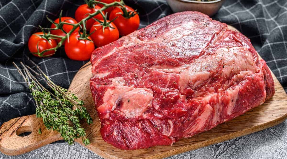 A chuck eye roast on a cutting board, in front of some herbs and tomatoes.