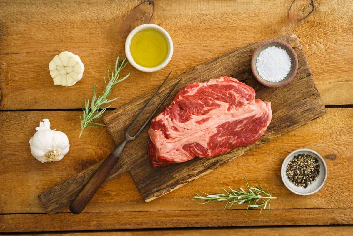 chuck eye steak on a wooden cutting board, with ramekins of oil and spi.