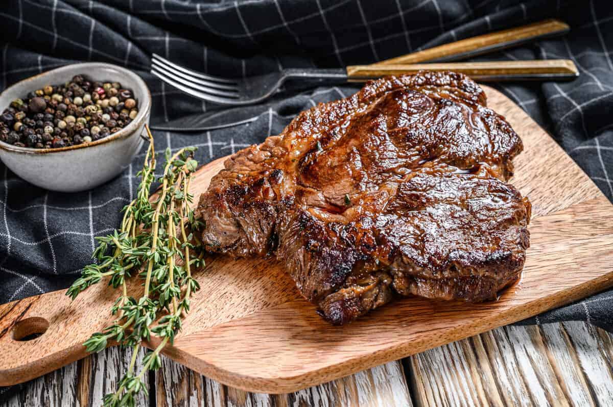 grilled chuck eye steak on a wooden paddle type board with a ramekin of salt and pep.