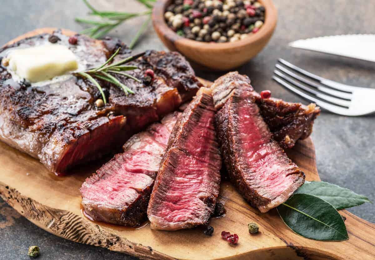 grilled and sliced ribeye steak on a cutting board with peppercorns and he.