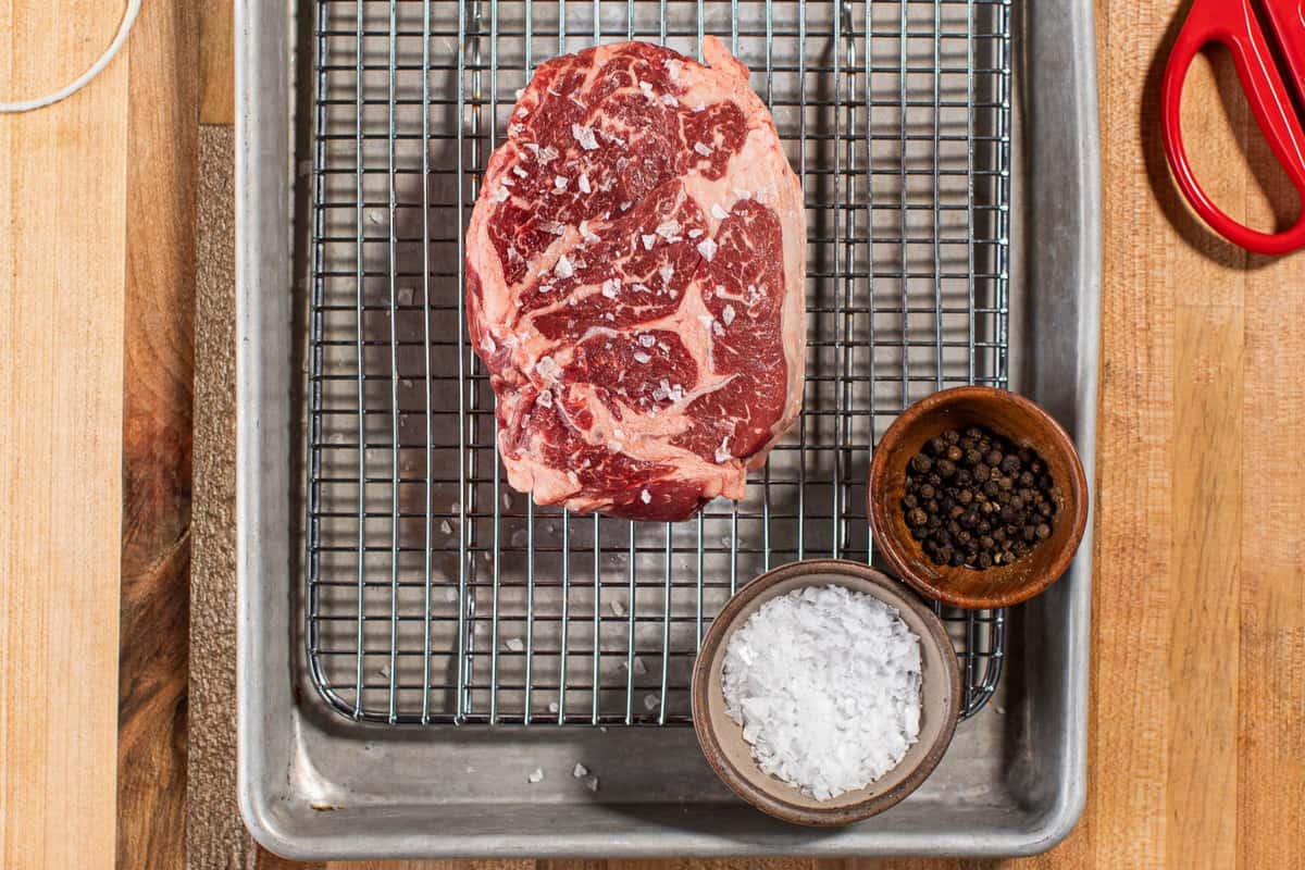 porter road chuck eye steak on a cooling rack, on a baking tray, with pots of salt and peppercorns