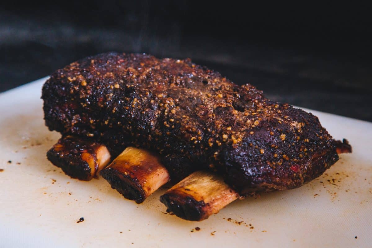 A rack of four smoked beef short ribs on a white cutting bo.