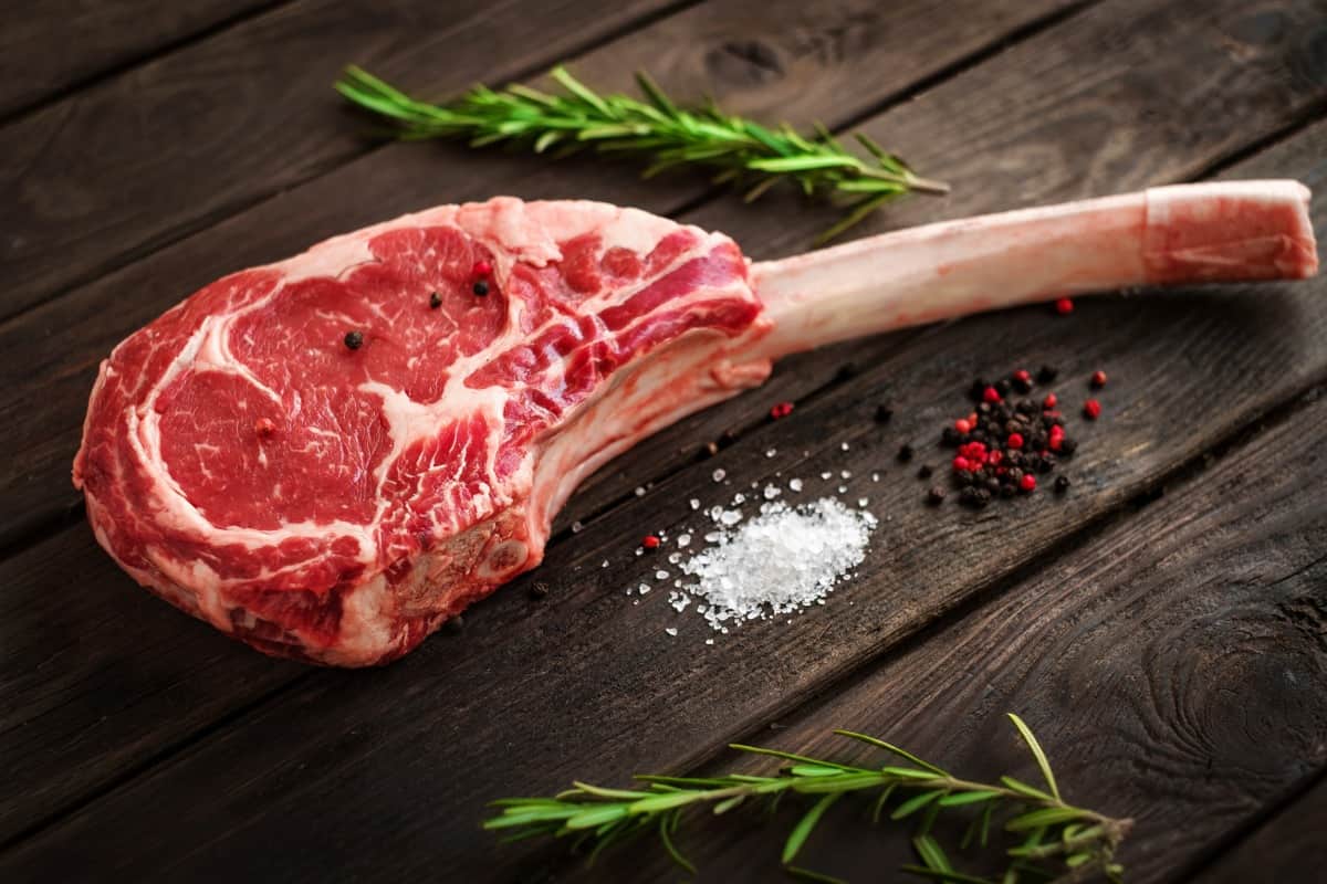 A tomahawk steak on a wooden table, next to small piles of slat and pepper, and two rosemary spr.