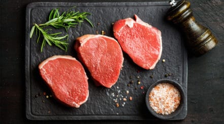 Three raw beef eye of round steaks with rosemary and rock salt on a dark surface.