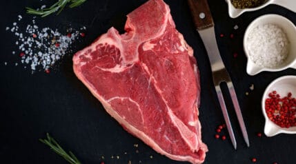 Raw porterhouse steak on a black background with some salt and spices in white dishes.