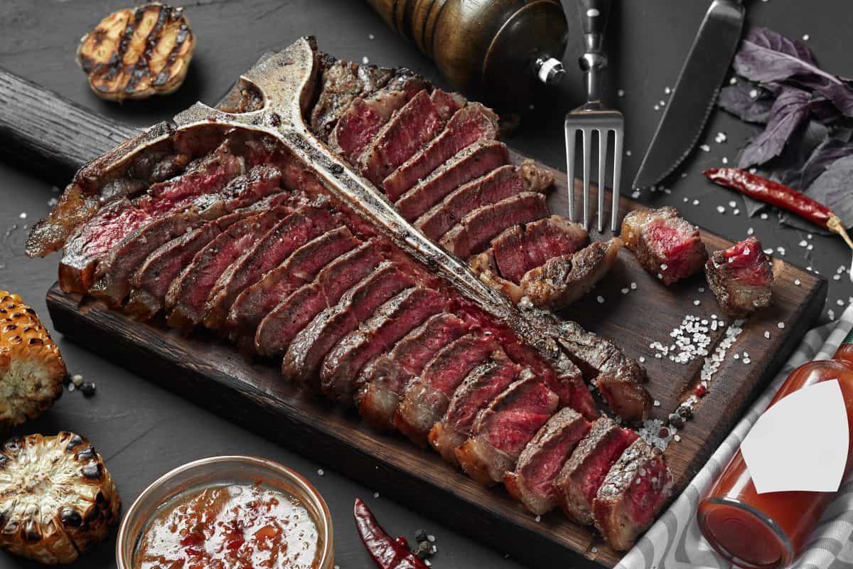 A grilled and sliced sliced porterhouse steak with grilled garlic halves and bottle of hot sauce.