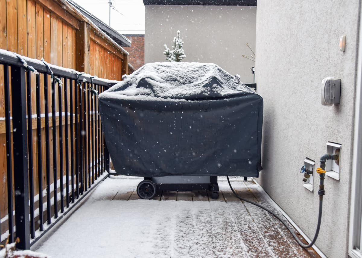 A gas grill on a balcony, under a cover that is being snowed .