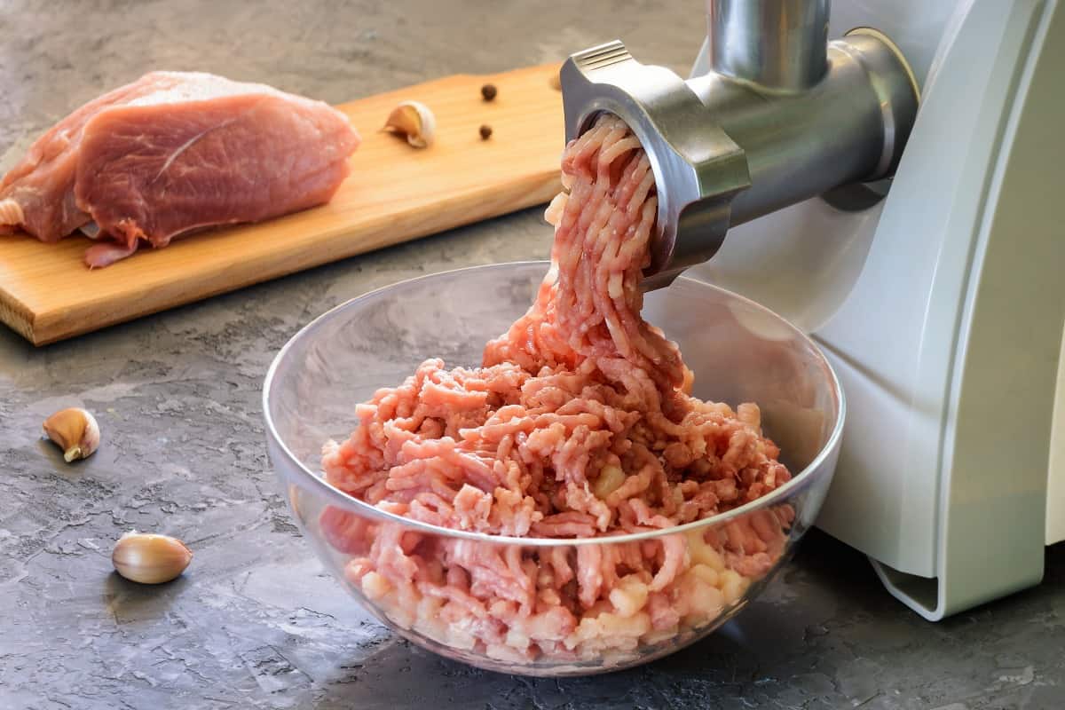 Ground meat coming out of a meat grinder, with a joint of meat and some garlic and peppercorns on a cutting board in the backgrou.