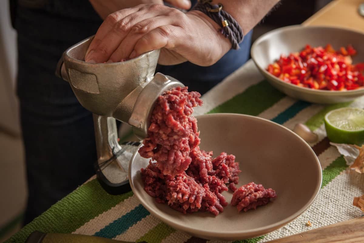 Mans hands forcing meat into a manual meat grinder, with mince coming out the end