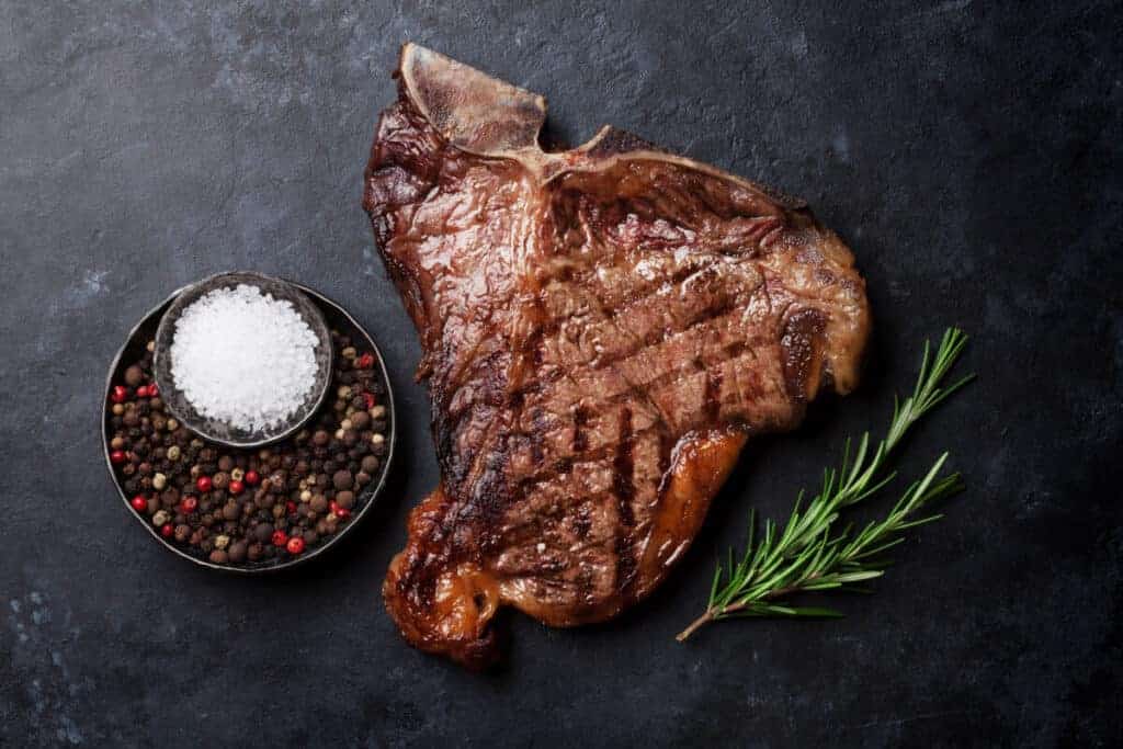 A grilled t-bone steak on a slate surface with salt and pepper p.