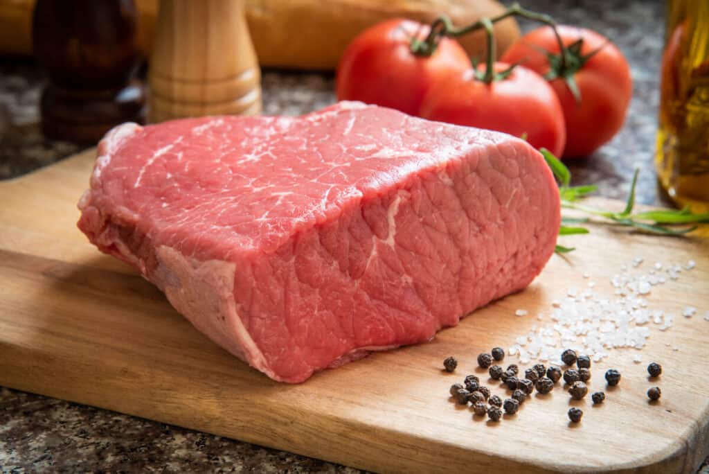 Uncooked bottom round roast joint, on a cutting board with a few black peppercorns in fr.