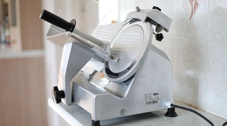 A silver commercial meat slicer on a granite kitchen work surface.