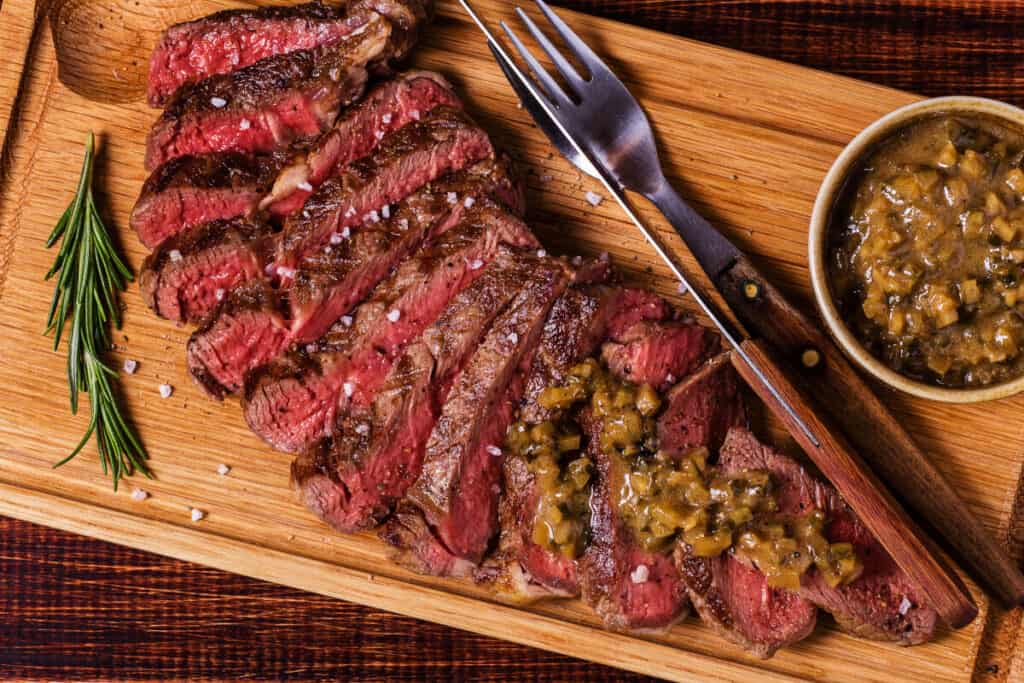 Grilled and sliced ribeye on a cutting board, with knife and fork and a mustard based sauce