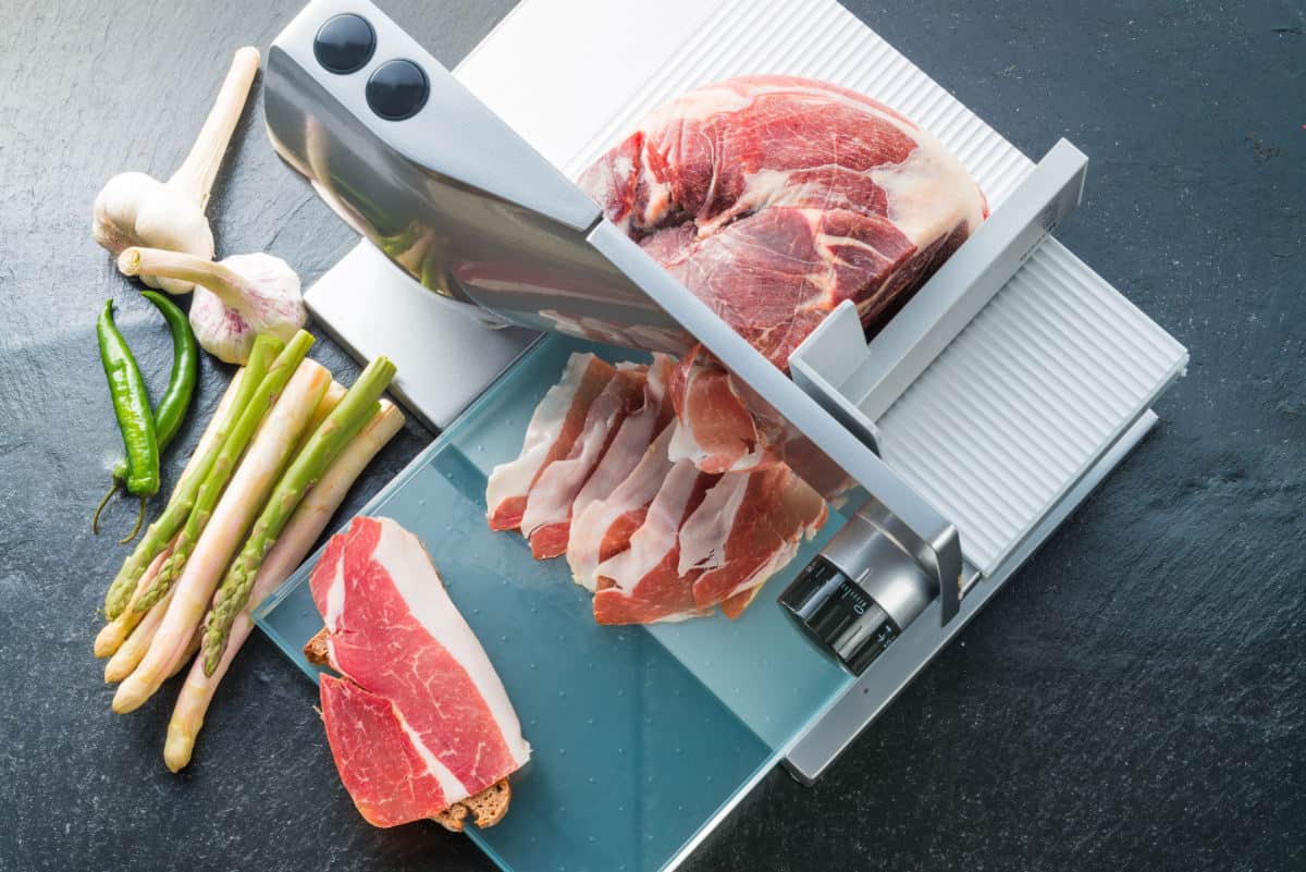 Bacon being sliced in a food slicer, with some garlic and spring onions in s.