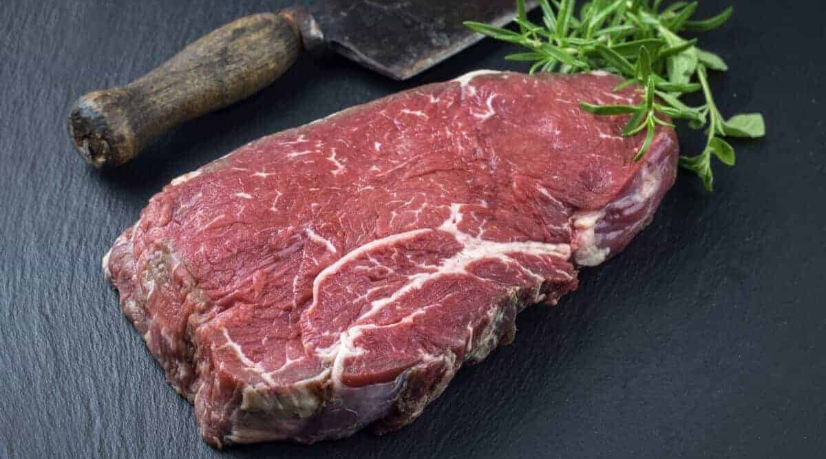 Raw bottom round steak, on a slate cutting board with rosemary and a butchers knife.